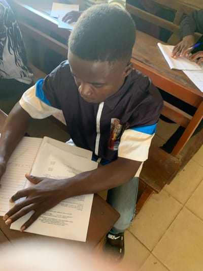 A young dark-skinned male with short dark hair is seated at a wooden desk in what appears to be an educational setting in Abobo, Nigeria. He is viewed from an elevated perspective, looking down at materials on the desk. He wears a collared, short-sleeved shirt featuring a dark blue or black torso with white vertical panels and light blue accents on the shoulders. His right sleeve exhibits a patterned design. He is also wearing light blue denim jeans and black Nike slide sandals.
His left hand holds open a spiral-bound notebook containing handwritten text, while his right hand rests upon a stack of white papers printed with black text. The individual appears to be engaged in studying or writing. To the right and behind him, a dark-skinned hand holding a blue pen is visible, positioned over white papers on an adjacent wooden desk, indicating the presence of at least one other person performing a similar activity. The floor is tiled with light-colored square tiles. Wooden desks and benches are arranged in rows.