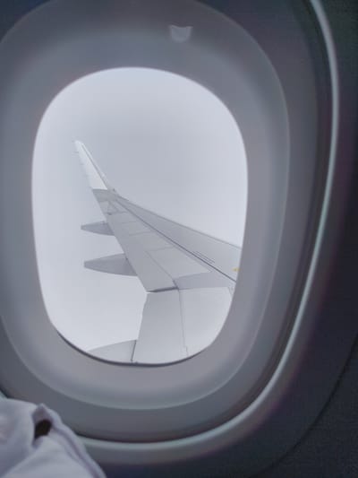 The image shows an airplane wing viewed through an airplane window. The wing is white and extends diagonally across the frame. The sky outside is overcast and gray. The interior of the airplane cabin is visible around the window, with a blurred white fabric in the foreground. The scene is consistent with being in an airplane, but there are no specific details in the image that can be used to determine the location in the city of Vinh Trach, Vietnam.