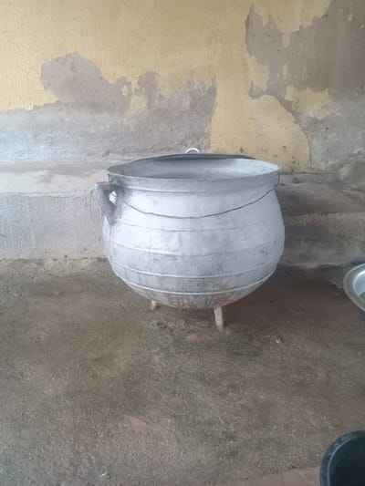 A large, weathered, aluminum cauldron sits on three metal legs on a dirt and concrete floor. The cauldron has a rounded body with horizontal ridges and two wire handles attached to the rim. A thin wire circles the upper portion of the cauldron. Behind the cauldron is a textured wall with peeling yellow and gray paint. To the right of the cauldron, partially visible, is a shallow metal basin. The scene appears to be an outdoor area, possibly a courtyard or cooking space, in Gbaza, Nigeria.