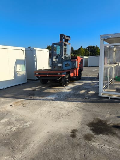 In the Municipal Unit of Inofyta, Greece, a front-end loader, painted in red and blue, is parked on a paved surface. The loader has a clear glass cabin, and the name "ISOBOX" is visible on its side. Behind the loader, several white portable structures are lined up, resembling modular buildings or containers. In the distance, a line of trees forms the horizon against a clear blue sky. The ground is a mix of concrete and gravel, with some oil stains and scattered debris. To the right of the loader, a white, open-sided structure with shelves is partially visible, containing green bins. The scene appears to be an outdoor storage or construction area.