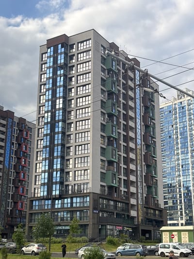 A medium-wide, eye-level shot captures a modern residential building complex in the city of Sennitsa, Belarus, under a cloudy sky. The primary focus is a tall, multi-story building with a facade of light gray concrete, dark brown accents, and large, reflective blue-tinted windows. The building features numerous balconies, some enclosed with glass and others open, with dark railings. Green rectangular modules are integrated into the facade, creating a staggered effect on some levels. Power lines crisscross the upper portion of the frame.
To the left, a similar, though slightly shorter, building is visible, displaying red and dark brown colored sections. To the right, a much taller building with a predominantly blue glass facade stands out.
At the base of the main building, commercial storefronts with large windows and signage are visible. A few trees and manicured green lawns with bushes line the sidewalk and street in front of the buildings. Several cars, including a silver SUV, a white van, and other dark-colored vehicles, are parked along the street. A lone individual in dark clothing walks on the sidewalk in the foreground. The overall impression is one of a contemporary urban or suburban residential development.