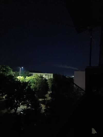 The image depicts a nighttime scene in Ladyzhyn, Ukraine, overlooking trees and buildings under a dark blue sky. A streetlamp illuminates a portion of the foliage, casting a yellowish glow. In the background, a light-colored building with visible windows stands behind a dense cluster of trees. To the right, a dark structure partially obscures the view, with a white wall visible beyond it. A faint plume of smoke rises in the distance. The foreground is dominated by silhouetted trees, rendering the lower portion of the image very dark. There are no people or discernible actions captured in the scene.