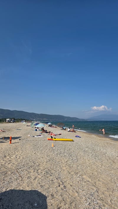 The image captures a sunny day at a pebble and sand beach in Ασπροβάλτα, Greece. The sky is bright blue with faint high-altitude clouds. A range of green, tree-covered mountains dominates the background, with distant peaks and a large white cumulus cloud visible on the far right. The sea is a dark blue-green, exhibiting small waves breaking gently at the shore.
Approximately 20-30 individuals are present on the beach and in the water. Many are seen sunbathing on towels or directly on the sand, while others are seated under various beach umbrellas (blue, green, and dark grey). Two individuals are partially submerged in the water on the right side, one appearing to enter and another further out.
Key objects on the beach include a prominent yellow inflatable object, possibly a paddleboard or raft, with black fins, positioned in the mid-ground. Two small orange traffic cones are also visible on the sand. A blue pair of sandals rests near the yellow inflatable. In the far left background, a white recreational vehicle (RV) and other vehicles are parked near the base of the mountains. A large, dark shadow from an unseen object extends into the lower left foreground.