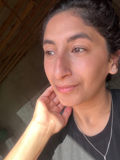 The image shows a close-up, head-and-shoulders shot of a woman with dark hair pulled back. She is wearing a black t-shirt and a silver necklace with a pendant. Her left hand is resting on her chin and cheek. She is wearing a small, curved earring with small stones on her right ear. Her skin has a natural, dewy glow, and her lips are slightly parted. The background is out of focus, but appears to be an indoor space with wooden beams and a window casting light and shadows. There is no indication of the city of Dondukovskaya, Russia, in the image.