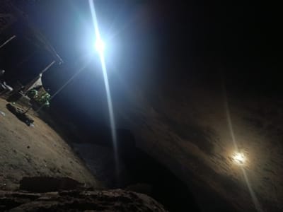 The image depicts an outdoor scene at night, likely in Masaka, Nigeria, illuminated by artificial lighting. The primary subjects are two bright light sources, one in the upper left and another in the middle right of the frame, casting long beams and creating lens flares. The foreground is dominated by dark, rough terrain, possibly soil and rocks, with some debris scattered on the ground.
In the background, to the left, a small structure or stall is visible, with some items, possibly produce or goods, arranged on it. The overall scene suggests a dimly lit, possibly rural or semi-urban environment at night. The bright lights indicate the use of artificial illumination, possibly for security, work, or a gathering.
The time of day is clearly night due to the darkness and the artificial lights. No specific weather conditions are discernible. The terrain and the presence of a simple stall suggest a location that is not highly developed. The mood is somewhat mysterious and utilitarian due to the stark lighting and the dark surroundings. There is no visible text in the image.
