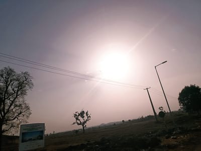 The image presents an outdoor scene in Rafin Pa, Nigeria. The sun is high in the sky, creating a bright, somewhat hazy atmosphere. Power lines stretch across the upper portion of the image, supported by utility poles. A street light is positioned to the right. Trees of varying sizes dot the landscape, silhouetted against the bright sky. In the lower left corner, there is a poster with the words "Groundbreaking Ceremony" on it. The environment appears to be a somewhat barren field or open area.