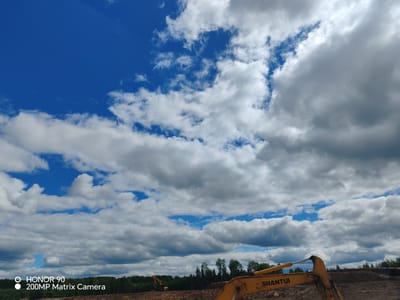 The image depicts a bright, cloudy day with a vast expanse of blue sky dominating the upper portion. White and gray clouds are scattered across the sky, with patches of blue visible between them. In the lower half of the image, a construction site is visible. A yellow excavator with the logo "SHANTUI" is prominently featured in the foreground, its arm extended. In the background, another yellow excavator can be seen in the distance. A dense line of green trees forms the horizon. The ground appears to be rough terrain, likely soil and debris from the construction activity. The image also contains a watermark in the lower-left corner that reads "HONOR 90 200MP Matrix Camera". The location is identified as Murashi, Russia.