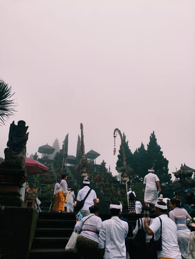 The image captures a scene at Pura Penataran Agung Lempuyang, a Hindu temple located in Rendang, Indonesia. The environment is misty, obscuring the background. Several people are present, many ascending the temple steps. The people are largely dressed in traditional Balinese attire, consisting of white shirts or blouses and sarongs. Many are wearing white headbands. There are some individuals carrying bags, and some of them are walking on the stairs. Temple structures are visible, along with decorative elements, and some trees in the background. There is also a red umbrella. The overall mood is serene.