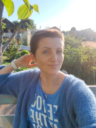 A woman with short brown hair and blue eyes is posing for a selfie outdoors, with her hand resting behind her ear. She is wearing a blue knitted cardigan over a blue t-shirt with white text. She has a white watch on her left wrist and a delicate silver necklace with a dark pendant. The background suggests a rural setting, possibly a dacha or backyard, with buildings, trees, and a greenhouse visible. The sun is shining, casting a warm glow and creating lens flare. The woman appears relaxed and is smiling gently at the camera. The location is identified as Дзямідавіцкі сельскі Савет, Belarus.
