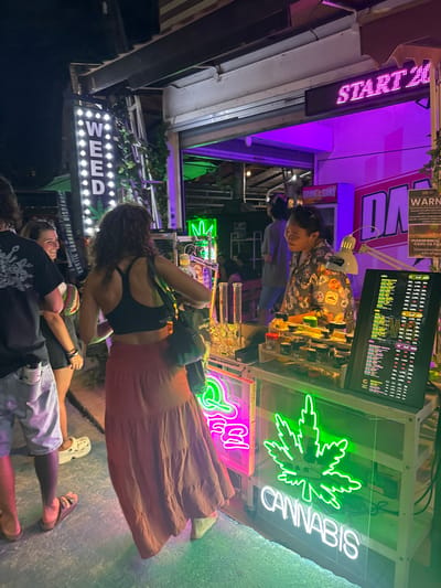 The image displays a nighttime scene at an outdoor cannabis dispensary on a street in Ban Ko Li Pe, Thailand. In the foreground, a woman with curly brown hair, wearing a black sports bra and a long, rust-orange tiered skirt, stands with her back partially turned, facing the dispensary counter. A black shoulder bag hangs from her left shoulder. To her immediate left, partially visible, is a man in a black t-shirt, light-colored shorts, and dark sandals, with a small white container clipped to his pocket. Behind him, a second woman, wearing dark attire and light-colored sandals, smiles and looks towards the man.
Behind the counter, a vendor with short dark hair, wearing a patterned short-sleeved shirt, smiles and looks towards the woman in the foreground. The white display counter features various products, including multiple small glass jars containing cannabis flower arranged on wooden risers, and two tall glass bongs or water pipes. An illuminated menu board on the right side of the counter lists cannabis strains and prices, indicating "1,000 THB" and "800 THB" among others. A white adjustable desk lamp is positioned on the counter. Below the counter, a prominent green neon sign depicts a cannabis leaf with the word "CANNABIS" in white neon below it, alongside a partially visible pink neon sign.
The establishment's exterior includes a tall, vertical white LED sign spelling "WEED" with a cannabis leaf graphic, and text reading "EXOTIC CANNABIS DISPENSARY". Above the entrance, a red LED ticker displays "START 20". A black and yellow "WARNING" sign with text in Thai and English, including "Please show ID Card", is visible to the right of the entrance. The interior of the shop is illuminated with purple and green ambient lighting. The ground is a light-colored, possibly wet or reflective, paved surface.