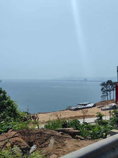 The image depicts a coastal scene in Hải Vân Ward, Vietnam, viewed from an elevated vantage point on a sunny day. A vast expanse of calm, blue water dominates the middle ground, extending to a hazy horizon where distant landmasses and faint urban buildings are visible. The sky above is a light, clear blue, with a distinct light ray or lens flare originating from the upper right quadrant.
In the immediate foreground, dense green foliage and exposed brown soil with rocks occupy the left side. Black cables are visible snaking across the dirt. To the right of this, a cleared, lighter brown dirt area slopes down towards the water. On this cleared area, multiple stacks of grey concrete pipes are observed, some partially covered with white tarpaulins or sheeting. Sparse green vegetation, including small shrubs and trees, is scattered across this dirt patch. A narrow, dark-colored path or road runs horizontally across the cleared area.
On the right side of the image, near the water's edge on the elevated land, several slender evergreen trees stand. A metal guardrail, likely belonging to a road, is present in the lower right corner, partially obscuring a red and white triangular road sign. No individuals or discernible activities are present within the scene.