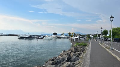 This media file depicts a scenic view of a harbor in Peschiera del Garda, Italy. The scene is set on a bright, partially cloudy day, suggesting it is likely daytime. The harbor is filled with various yachts and boats docked along a pier. In the foreground, a rocky shoreline leads to the calm, blue water of the lake.

To the right of the harbor, a paved walkway runs alongside a road, bordered by trees and lampposts. Several cars are parked along the roadside. Buildings, likely part of the town, are visible in the background, interspersed with lush green trees and distant mountains that fade into a hazy blue. The overall atmosphere is peaceful and serene, conveying a sense of a typical summer day at a lakeside town.

Visible text includes "POLIZIA PROVINCIALE" on one of the boats, indicating a police vessel. There are also parking signs visible along the road. The presence of yachts and the well-maintained walkway and road suggest a popular tourist destination or a well-established marina.