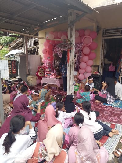 A group of children and women are seated on mats, attending an outdoor celebration. Pink decorations, including balloons and a backdrop, adorn the area. A table with a tiered cake and other treats stands nearby. Some attendees are wearing headbands, while others are in hijabs. There is a stuffed pink bear behind the table. One woman is standing and seems to be helping with food.