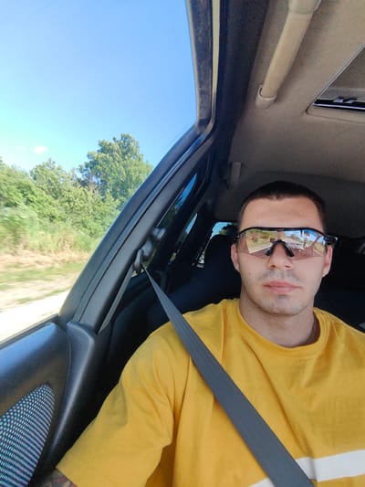 A man wearing a yellow t-shirt and sunglasses is sitting in the driver's seat of a car. The car appears to be moving, as indicated by the motion blur of the trees and landscape outside the window. The man is looking directly at the camera, and his sunglasses reflect the road ahead. The interior of the car is visible, including the dashboard, seatbelt, and part of the ceiling. The scene is captured on a sunny day, with a clear blue sky. The location is the road outside of Stefanovo, Bulgaria, a small village nestled in a rural area.
