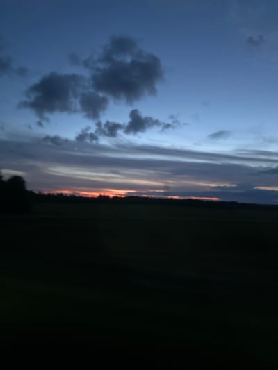 The image depicts a rural landscape at dusk in the vicinity of Грозаўскі сельскі Савет, Belarus. The sky is a deep blue, transitioning to lighter shades near the horizon, with streaks of orange and pink from the setting sun visible through scattered clouds. The clouds are dark and textured, adding depth to the sky. Below the sky, a dark silhouette of trees lines the horizon, suggesting a wooded area. In the foreground, a vast, dark expanse of what appears to be a field or open land occupies the lower half of the image, rendered almost entirely in shadow due to the low light conditions. There are no people or discernible objects visible in the frame, and the overall scene is one of natural, undisturbed twilight in a rural setting.