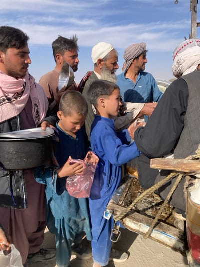 The image captures an outdoor scene in Chaman City Tehsil, Pakistan, under a clear blue sky with sparse white clouds. A group of approximately seven male individuals, ranging from young boys to elderly men, is gathered, appearing to queue or participate in a distribution event.
In the foreground on the left, a young man, wearing a light pink shawl over a dark jacket and light-colored traditional clothing, holds a black pot with a metal plate resting on top. Next to him, a young boy in a light blue tunic looks downwards, clutching a pink plastic bag. To the right of this boy, another young boy, dressed in a bright blue tunic, extends his hands forward, seemingly receiving an item from an individual or station located to the right and partially outside the frame.
Behind these two boys, an adult man with dark hair, wearing a brown shirt, holds a metal bowl. To his right, an older man with a long white beard and a white cap is visible. Further to the right, another adult male with a grey turban and blue shirt extends his arm forward, engaged in the act of receiving. On the far right, partially obscured, an individual in dark clothing and a white and patterned turban stands near a wooden structure featuring ropes, which appears to be part of a makeshift dispensing station or cart.
The ground is light-colored and dusty, indicating an arid environment. Strong shadows are cast, suggesting bright overhead sunlight. A light-colored vehicle is faintly visible in the distant background on the right.