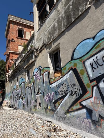 An exterior low-angle shot shows an abandoned building in Meljine, Montenegro. The building has exposed brick sections, deteriorating concrete, and multiple windows, some broken or missing. Graffiti covers the walls. The ground is a mix of rubble and debris. The sky is clear blue.