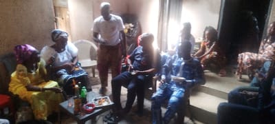 The scene depicts an indoor social gathering of approximately eight to ten individuals in Ohafia, Nigeria. In the foreground, a small, dark rectangular table holds multiple plastic bottles of beverages, several cups, and at least two small plates containing snacks.
On the left, two women are seated. The leftmost woman wears a yellow garment and a purple headwrap, with visible jewelry including a necklace and bracelets. To her right, an older woman wears a white top, a dark patterned skirt, and a dark headwrap; she appears to be holding a small object in her hand.
Near the center-left, a man stands wearing a white short-sleeved t-shirt and light brown or patterned trousers. To his right, a man is seated in a dark short-sleeved shirt and dark trousers, holding a small, light-colored object. Further to the right, another man is seated, dressed in a blue and white patterned long-sleeved outfit, holding a light-colored plate with contents.
Additional individuals are visible in the mid-ground and background, some seated on chairs or on the floor/steps, and some less distinct due to lighting and depth. The walls are light brown or beige, appearing to be textured or unpainted. A doorway is visible in the background, leading to a darker space. The floor is concrete, with visible steps on the right side of the frame. The overall impression is a casual gathering with refreshments.