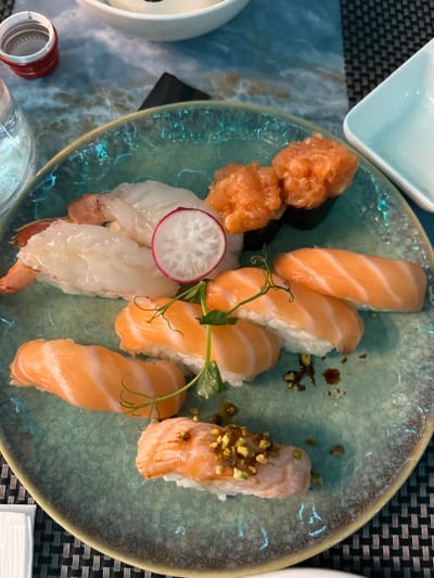 An overhead shot captures a visually appealing assortment of sushi served on a round, light blue-green ceramic plate with a textured, rustic rim. The main subjects are various types of sushi.

Prominently featured are multiple pieces of salmon nigiri. Some pieces are plain, showcasing the fresh orange salmon draped over white rice. Others are garnished with delicate green pea shoots. Additionally, at least two pieces of salmon nigiri are topped with a glossy sauce or glaze and sprinkled with chopped pistachios, adding a unique texture and color.

Towards the bottom left, there are two pieces of shrimp (ebi) nigiri, with the pinkish-white shrimp positioned on rice. To the far left, two distinct pieces of what appear to be salmon tartare or spicy salmon are presented on small bases, possibly rice or seaweed, forming a gunkan maki style. A thin, decorative slice of radish with a pink edge adds a fresh accent to this section of the plate.

The plate rests on a table with a blue and white marbled pattern, which contrasts with a dark gray woven placemat partially visible on the right. In the top left corner, a small, square light-blue dish, likely for soy sauce, is partially visible. Other glimpses of tableware include a white bowl and a red bottle cap in the bottom left, and a clear glass partially seen at the very bottom.

The image depicts a meal, likely enjoyed in a restaurant or at home in Basiglio, Italy, given the context. The lighting suggests an indoor setting, but the specific time of day, weather, or emotions are not discernible. There is no visible text in the image.