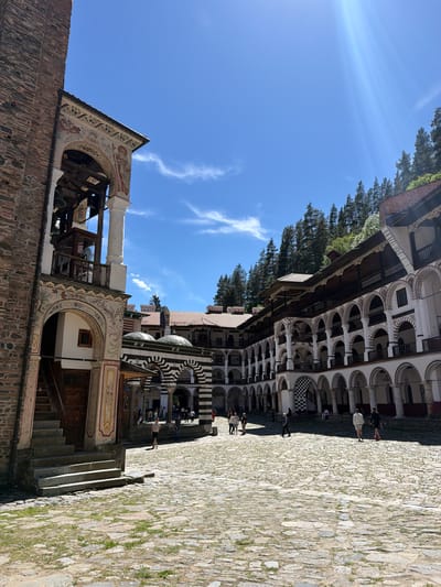 The image shows a portion of the Rila Monastery in Rilski manastir, Bulgaria. It depicts a courtyard paved with stone. A multi-storied building featuring arched colonnades and painted facades is visible, along with part of what seems to be a bell tower decorated with frescoes and columns. The sky is blue and partly cloudy. There are some people visible in the background, but their actions are not clear. Some trees are also present in the background.