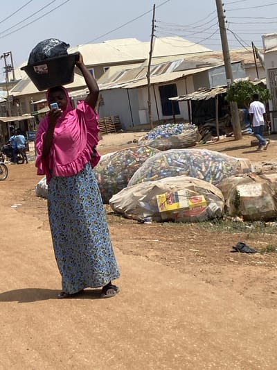 This image depicts a sunlit, unpaved street scene in Baganakwo, Nigeria. The central figure is a woman positioned slightly left of center, walking towards the viewer's left. She wears a vibrant pink head covering that extends over her shoulders, a matching pink top with ruffled sleeves, and a long blue skirt with a floral pattern. She carries a black plastic basin containing dark material on her head, stabilized by her raised right hand. Simultaneously, she holds a light-colored smartphone in her left hand, viewing its screen.
To her right, a collection of large, translucent white bags are stacked on the dirt ground, filled with various recyclables, predominantly plastic bottles and aluminum cans. One bag contains cardboard boxes. Behind these bags, a rudimentary structure with a corrugated metal roof is visible. Further in the background, a man in a white t-shirt and blue jeans walks away from the viewer towards the right. Several single-story buildings with corrugated metal roofs and light-colored walls line the street in the distance.
To the left of the central figure, a utility pole with multiple overhead wires stands. Two individuals are seated on a motorcycle parked near a building in the mid-ground. A partially visible sign displaying "BetK" is on a building in the background. The ground is reddish-brown dirt, showing texture and dust, with distinct shadows indicating strong overhead sunlight.