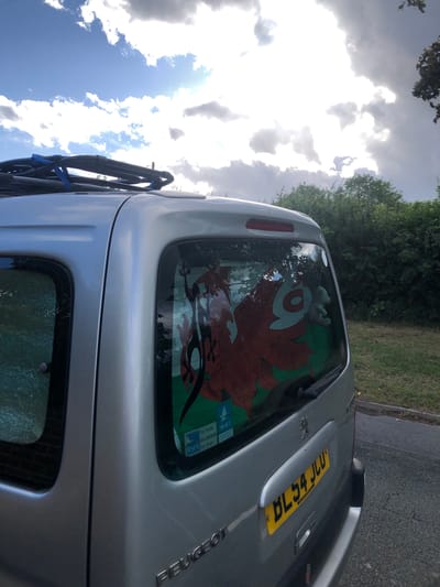 A silver Peugeot Partner van, license plate BL54 JCU, is parked on a paved road next to a grassy verge with trees in the background. The van has a roof rack and a sticker of the Welsh dragon on the rear window. There are two smaller stickers below the dragon sticker, one with the RSPB logo and text that appears to say "for birds, for people, for ever." The sky is cloudy with patches of blue. The location is the city of Shotton, United Kingdom.