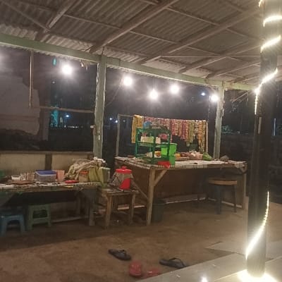The image depicts an unoccupied, semi-enclosed vendor stall illuminated at night in Ketowan, Indonesia. The structure features a corrugated metal roof supported by a skeletal framework of poles, from which several bare incandescent light bulbs are suspended, providing primary illumination. A vertical support pole on the far right is wrapped with a spiral string of white LED lights. The ground beneath the stall appears to be packed earth or concrete.

On the left, a rectangular wooden table holds various items including a blue plastic container, multiple cups, small bowls, and fabric-wrapped bundles. Two low plastic stools, one blue and one green, are positioned underneath this table.

Central to the scene is a larger wooden counter or display stall. Behind this counter, numerous pre-packaged snack items and instant noodles are suspended in rows from a horizontal line, indicating retail display. On the counter itself, a green, two-tiered plastic shelving unit holds several items, possibly containers or utensils. Miscellaneous goods, potentially produce or food items, are arranged on the counter surface. A dark, round-seated stool and a dark waste bin are situated beneath the counter.

Adjacent to the left side of the main counter, a red insulated beverage dispenser rests on a small, light-colored wooden stool. On the ground in the immediate foreground, two pairs of sandals are present: one pair of black flip-flops and one pair of red flip-flops. The background is dark, indicating nighttime, with faint distant lights visible. No individuals are present in the scene.