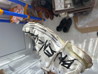 A human hand with light skin tone is holding a single, well-worn white Balenciaga Runner sneaker in an indoor, domestic setting. The sneaker features mesh and layered synthetic materials, with distinctive black, crudely applied, abstract, or character-like markings on its white upper panels. The word "RUNNER" is visible near the top of the tongue, and "BALENCIAGA" is on a side strap. Cream-colored laces are loose. The shoe exhibits signs of dirt and wear, particularly on the sole and midsole, which has a translucent yellowish hue. In the blurry background to the left, a blue and yellow plastic shoe rack holds several pairs of shoes, including pink children's sneakers. To the right, a dark brown multi-tiered shoe rack contains various adult shoes, including dark boots. Two pairs of dark slip-on shoes rest on the light-colored floor beneath this rack. Linear shadows from a light source, likely a window, are cast across a white surface in the lower left portion of the image. The specific city of Polkovnik Diakovo, Bulgaria, cannot be visually confirmed from the provided image.