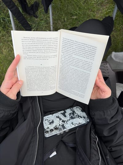 Here is a description of the image:

A person is seated, likely outdoors, reading a book. They are wearing a black jacket and black pants. The book is open, displaying text in Russian, and is positioned in front of them. A cell phone in a clear dragon patterned case is visible. The person is holding the book with both hands. The environment appears to be a mix of natural and constructed elements.