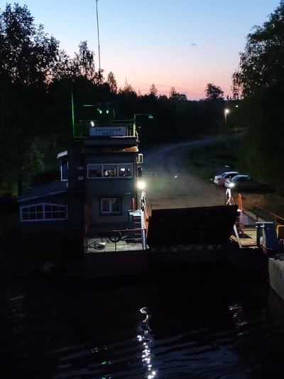 A nighttime view of the Annensky Most ferry crossing, Russia, shows a two-story control tower with a sign reading "ANNENSKIY MOST" on the top level. Green lights illuminate the sign and tower roof. The tower appears to be constructed of brick or stone, with windows on both floors. A spotlight shines from the tower onto the ferry deck. The ferry itself features a large ramp. Two cars are parked on the land side of the ferry crossing. The sky transitions from blue to pink near the horizon, indicating either sunset or sunrise. Trees line the background. The water in the foreground reflects light, with ripples visible on its surface.