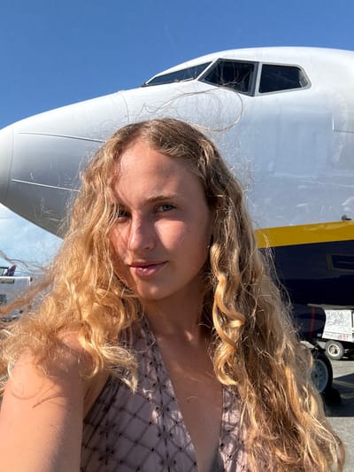 The image shows a close-up, selfie-style shot of a young woman with long, blonde, curly hair. She is positioned in front of the nose of a large aircraft, with the blue sky visible behind it. The woman's face is turned towards the camera, and she has a slight smile. Sunlight illuminates her face, creating highlights and shadows, suggesting it is daytime.
The aircraft appears to be a passenger jet, with its distinctive white fuselage and windows visible. A portion of the yellow and dark blue livery of the aircraft is also visible, hinting at an airline's branding. In the background, a wheel and part of the landing gear of the aircraft are discernible, along with some equipment or structures on the tarmac. The overall setting appears to be an airport or an area where aircraft are parked. The image captures a casual moment, possibly before or after a flight, with the woman seemingly enjoying the sunshine. The presence of the aircraft and the open sky suggest travel or a departure. No visible text is clearly legible within the frame.