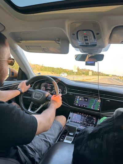 The image displays the interior of an Audi SUV, likely a Q7 or Q8, from the perspective of a front passenger, looking forward towards the driver and the road. The scene is illuminated by bright daylight, suggesting late afternoon or early morning due to the warm light tones.
A male driver, with short dark hair and wearing sunglasses, a black short-sleeved t-shirt, and grey cargo shorts, is seated behind the steering wheel, which bears the Audi logo. He grips the wheel with both hands. A dark-strapped watch is visible on his left wrist. The driver is wearing a seatbelt, visible across his lap.
The car's digital instrument cluster displays "Consumption," the gear "D8," an odometer reading of "139633 km," and a range of "553.4 km." The central infotainment screen shows a navigation map with a blue route line. The display indicates "30 km" (to an unspecified point), "22:20 arrival," a "3:18 drive time," and "248 km" remaining to the destination, which is identified as "Thessaloniki." A road labeled "A3" is visible on the map. Below this, a separate touch screen manages climate control, showing "24.0" degrees Celsius for the left zone and "18.0" degrees Celsius for the right zone, along with "AUTO" settings and seat ventilation controls. The electronic gear selector is in "P" (Park). An overhead console features controls for interior lighting and a button with a red illuminated indicator. A sunroof is partially visible at the top of the frame. A rear-view mirror is mounted centrally, with a small electronic device, possibly a dash camera, attached below it by a cable.
The vehicle is on a multi-lane highway with an asphalt surface. The surrounding landscape consists of hills with sparse, dry vegetation. A white car is visible in the distance on the highway. A white road sign with a dark border is present on the right shoulder, but its text is illegible. The windshield has visible water spots or residue, slightly obscuring the view. The location is stated to be Valkovo, Bulgaria; however, the navigation system in the vehicle indicates a destination of Thessaloniki, 248 km away, which is geographically inconsistent with a starting point in Valkovo, Bulgaria.