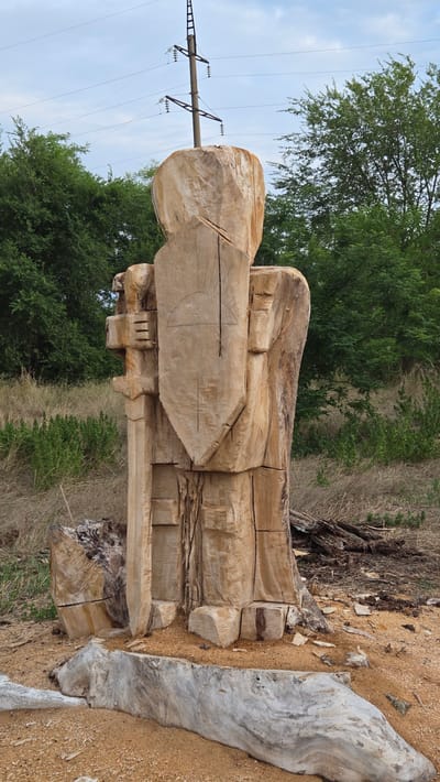 A large, unfinished wooden sculpture resembling a knight or warrior stands outdoors. The sculpture is carved from a tree trunk and features a shield on its chest and a sword-like object in its left hand. The raw wood shows visible tool marks. In the background, there are trees, dry grass, and a power line pole with wires. The ground around the sculpture is covered with sawdust and loose wood. A light-colored, weathered log or stone structure is in the foreground. This scene is located in Nezavertaylovka, Moldova.
