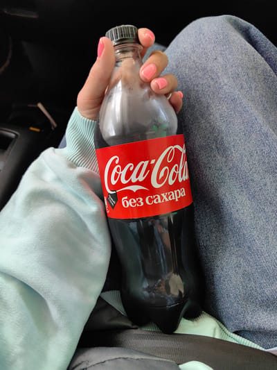 The image shows a close-up of a person holding a bottle of Coca-Cola Zero Sugar. The bottle has a red label with the Coca-Cola logo and text in Russian that translates to "without sugar". The person's hand, with pink nail polish, is visible holding the bottle. They are wearing a light blue sweatshirt and sitting on a surface covered with blue denim fabric. The background is dark and out of focus, suggesting an indoor or dimly lit environment. There are no visible landmarks or indicators that would specify the location within Фаніпальскі сельскі Савет, Belarus. The scene appears to be a casual moment, possibly while traveling or relaxing.