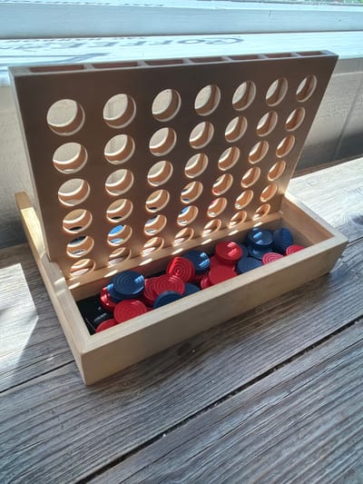 This media file depicts a close-up, high-angle view of a Connect Four game. The game board, made of light-colored wood, is open and angled upwards, revealing a wooden box filled with red and blue game pieces. The board has rows of circular holes through which the game pieces would fall during play. The pieces themselves are circular and appear to be made of plastic or wood, with visible ridges. Some pieces are already in the box, while others are scattered within it. The background is composed of weathered wooden planks, suggesting an outdoor or rustic setting. Sunlight streams in from the left, casting shadows on the wooden planks and the game board, indicating it's daytime, likely morning or afternoon. There are no people visible in the image. The overall scene suggests a moment of pause in a game, or the storage of game pieces. No text is visible in the image. The setting could be a porch, patio, or a room with wooden furniture.
