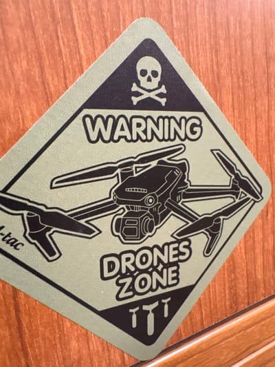 A diamond-shaped sticker with rounded corners is affixed to a reddish-brown wooden surface exhibiting horizontal grain. The sticker features a sage green background with black elements. Centered at the top is a skull and crossbones symbol, followed by the bold capitalized text "WARNING." Below this, a detailed black outline of a quadcopter drone, resembling a DJI Mavic series with an underslung camera, dominates the central area. The text "DRONES ZONE" is positioned beneath the drone, and three stylized black silhouettes resembling stick grenades or small bombs are at the bottom. A partial "l-tac" inscription is visible along the left edge. The scene is brightly illuminated. The precise location within Richmond, United States, cannot be determined from the visible image data, but the item is adhered to an indoor-appearing wooden surface.