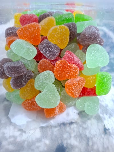 A close-up, overhead shot shows a pile of heart-shaped gummy candies in a clear plastic container. The candies are various colors, including green, orange, red, yellow, and purple. They have a granular, sugar-coated texture. The container appears to be partially empty, with a cloudy, white surface visible underneath the candies. A few stray hairs are also visible on the clear surface of the container. The location is Kutaglumpang, Indonesia.