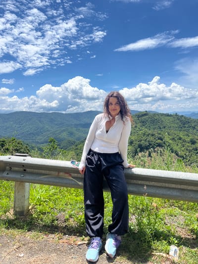 A woman with curly brown hair is posing for a photograph while leaning on a metal guardrail. She is wearing a white long-sleeved zip-up top with a black bra visible underneath, black baggy pants, and light blue and purple athletic shoes. Her left hand is resting on the guardrail, and she is looking directly at the camera. She has a light smile.
The background is a lush green mountainous landscape with a bright blue sky filled with scattered white clouds. The location appears to be a scenic overlook, possibly on a mountain road. There are green plants and grass growing alongside the guardrail and on the embankment. A pair of sunglasses can be seen resting on the guardrail near the woman's left hand.
While the image features a mountainous landscape and cloudy sky, the specific location of Son Dien, Vietnam, cannot be definitively determined from the visual information provided.
