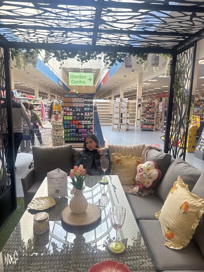 The image depicts the interior of a large retail space, possibly a department store or garden center, in Portadown, United Kingdom. A young woman with dark hair is seated on outdoor furniture, making a peace sign with her hand. The furniture, identified by a nearby sign as a "Florence Corner Set," includes a gray sofa with decorative pillows, a glass-topped table, and a chair with a gray cushion. On the table are decorative items such as a vase with tulips, a wooden slice, a plate, and glassware.

In the background, other shoppers are visible, and the store's merchandise is organized on shelves, including gift cards and seasonal items. A green sign directs customers to the "Garden Centre." The scene is partially framed by a decorative metal structure with a tree branch design, adding a unique visual element to the setting. The lighting is bright and diffused, suggesting a well-lit retail environment.