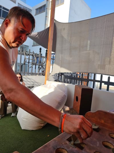 A man in Magdalena del Mar, Peru, is standing next to a wooden game board with round holes. He is inserting a flat metal piece into one of the holes. The man is wearing an orange bracelet and a white tank top. He has short, dark hair. Behind him is a large white cushion on green artificial turf. A woman wearing sunglasses is sitting behind the cushion. A metal railing and a white building are visible in the background. An awning provides shade.