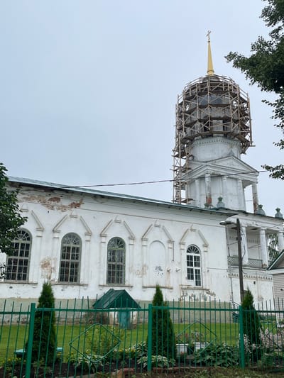 The image shows a white Orthodox church with a golden cross on top, situated in Kstinino, Russia. The church's dome is partially covered in scaffolding, suggesting it is undergoing renovation. The church has a long, white facade with arched windows that have green frames. A green metal fence encloses the front of the church, with manicured bushes and flowers visible behind it. The sky is overcast, and a few trees can be seen on the right side of the image. There are no people visible in the image.