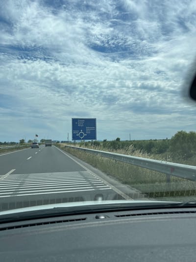 The image depicts a view from inside a vehicle, looking forward along a highway. The road is paved with multiple lanes, marked by white lines. A highway sign, blue with white lettering and a circular arrow diagram, is visible on the right side of the road. The sign indicates directions to "Pomorie," "Burgas," "Kableshkovo," and "Aheloy." The landscape beside the highway consists of dry grass and scattered trees under a cloudy sky. Two cars and a truck are on the highway ahead. A Bulgarian flag is planted on a pole to the left of the road.