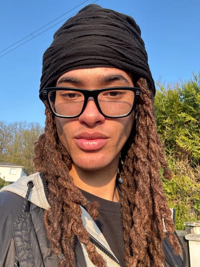 A close-up outdoor portrait depicts an individual of medium-brown complexion, looking directly at the camera. The subject wears a black textured headwrap, black-framed rectangular glasses with clear lenses, and has long, thick, dark brown dreadlocks extending down past the shoulders. Small black bands secure some of the dreadlocks. The individual is dressed in a dark inner garment and a dark gray outer jacket featuring lighter gray elements on the hood and a black zipper. Natural sunlight illuminates the scene from the right, creating highlights on the right side of the face and hair. The background, located in the city of Marcoussis, France, features a clear blue sky. To the left, a partially visible light-colored residential building with a dark roof stands behind trees with sparse foliage. To the right, dense green coniferous foliage is visible. A light-colored stone pillar is present in the lower right foreground. Faint power lines are discernible against the sky in the upper left.