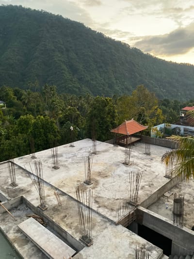 The image depicts an unfinished building structure on a rooftop in Sawan, Indonesia. The foreground is dominated by a concrete slab with numerous vertical rebar posts protruding upwards, indicating ongoing construction. Several sections of the concrete slab are at different levels, suggesting a multi-story building in progress. To the left, a large, flat concrete slab leans against a lower level.

In the mid-ground, a small open-air pavilion with a traditional Balinese tiled roof stands on the rooftop. Beyond the pavilion, more construction is visible, including unfinished walls and rebar. A palm frond hangs into the frame from the right side.

The background reveals a lush, densely forested mountain slope that fills the majority of the upper portion of the image. The sky above is partly cloudy, with the sun appearing to be setting or rising, casting a warm glow on the clouds and the upper ridges of the mountain. The environment suggests a tropical location. No people are visible in the image.