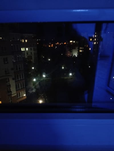 The image shows a nighttime view from a window. Buildings with illuminated windows are visible in the distance. There are also streetlights lining a park or open area. The location is possibly in Krykhivtsi, Ukraine, given the prompt. The immediate foreground consists of the window frame, which is blue. The overall impression is that of a residential area at night.