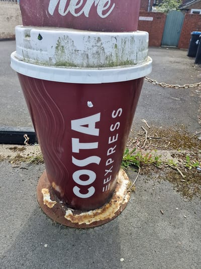 The image shows a Costa Express sign in the shape of a coffee cup, located in Pity Me, United Kingdom. The sign is maroon with white lettering that reads "COSTA EXPRESS". It has a white lid and is mounted on a rusty circular base. The sign appears weathered and has some moss growing on the lid. The ground is paved with asphalt. There is some vegetation growing around the base of the sign, and a chain is attached to the back of the sign. A red brick building and some bins are visible in the background.