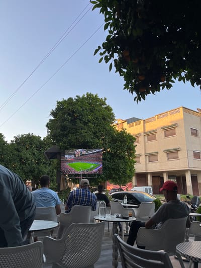 An outdoor scene at dusk depicts people watching a soccer match on a large screen set up amidst trees, possibly orange trees judging by the visible fruit. A building with beige stucco and brown-tinted windows is visible in the background. People are seated in white plastic chairs at tables, some with drinks on them. The sky is clear and blue. The scene appears to be a cafe or a public gathering space in El Hajeb, Morocco.