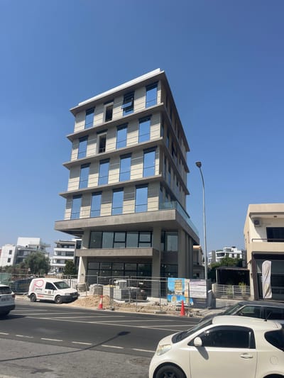 The image shows a modern, multi-story building under construction in Latsia, Cyprus. The building features a concrete exterior with large, reflective blue windows. Several floors are visible, with some windows open. There is a glass balcony on one of the upper levels. In front of the building, a construction site is fenced off, with piles of materials, construction equipment, and traffic cones. A white van with "prima KITCHENS" branding is parked on the side of the road. Other vehicles, including a white car and a dark-colored car, are on the road. Several other buildings, some of which appear to be residential, are visible in the background, along with trees and a clear blue sky. A lamppost stands near the construction site. The scene appears to be a street view on a sunny day.