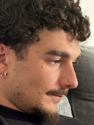 A close-up, profile image features a man with medium brown, curly hair and a neatly trimmed mustache and goatee. He is positioned towards the left of the frame, looking downwards and slightly to the right, suggesting focus or contemplation. His fair skin has several small moles or freckles on his cheek, forehead, and nose. A silver hoop earring is visible in his left ear. His visible eye appears to be light brown or hazel. He wears dark clothing, possibly a t-shirt or shirt, around his neck and shoulder. The background is softly blurred, revealing a grey, textured fabric, likely a blanket or part of a couch or chair, indicating an indoor setting. The lighting is soft and even, highlighting his facial features. No specific activity is clearly depicted, and no text is visible. The scene is set in Redondo Beach, United States.