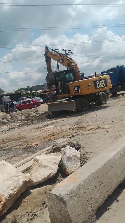 The image shows a construction site in Effurun, Nigeria. A yellow CAT excavator is the focal point, with its arm raised. Nearby are several large, irregular white rocks and a long, rectangular concrete block. In the background, there are people, vehicles including a red car and a blue truck, and buildings, suggesting an urban setting. Power lines are visible in the sky. The ground is uneven and appears to be dirt and gravel.